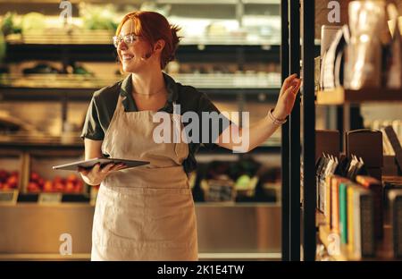 La propriétaire d'un magasin qui a réussi à sourire tout en tenant une tablette numérique dans son magasin d'alimentation. Une femme entrepreneure joyeuse qui a une petite entreprise dans le Th Banque D'Images