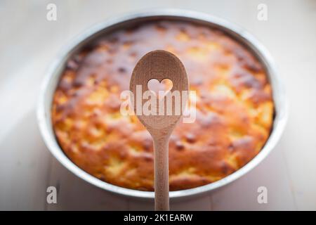 Cuillère en bois avec trou en forme de coeur, et tarte dans le fond sur table blanche. Concept cuit avec amour. Banque D'Images