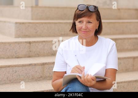 femme mature travaille à distance, l'enseignement supérieur pour les adultes, femme écrit et note dans un carnet assis sur les escaliers à l'extérieur, indépendant. Collège s Banque D'Images