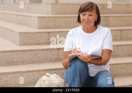 femme mature travaille à distance, l'enseignement supérieur pour les adultes, femme écrit et note dans un carnet assis sur les escaliers à l'extérieur, indépendant. Collège s Banque D'Images