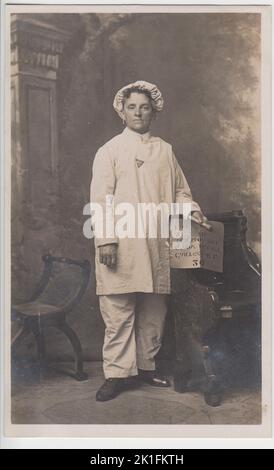 Portrait studio d'un travailleur de munitions de la première Guerre mondiale (un stencilor). Elle a été photographiée dans ses vêtements de travail de pantalon blanc, de chemise longue ou de salopette, et de la calotte de mob couvrant ses cheveux. Elle porte un badge triangulaire « On War Service » sur sa chemise et porte des boucles d'oreilles. Dans sa main gauche, elle tient un pinceau épais et un pochoir en papier qui aurait été utilisé dans l'usine pour marquer des boîtes de munitions Banque D'Images