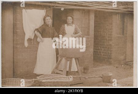 Journée de lavage dans le Bedfordshire d'Edwardian. Photo du début du 20th siècle de deux femmes vêtues de tabliers, debout près d'une grande baignoire de lavage qui a été équilibrée sur un tabouret. Deux paniers de linge en osier, un long bâton de transport et quelques seaux en métal sont sur le sol en face d'eux. Une chemise fraîchement lavée est accrochée à la ligne de lavage derrière elle. La photo porte le nom de deux photographes au dos - Ussher du 24, rue Battenberg, Bedford, et Lennox Gordon de Woburn Sands. Banque D'Images