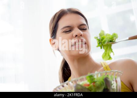 Jeune femme caucasienne assis sur un canapé en mangeant de la salade de légumes tout en se sentant dégoûtée et l'expression dégoûtée, femme émotion frustrée avec l'anorexie ve Banque D'Images