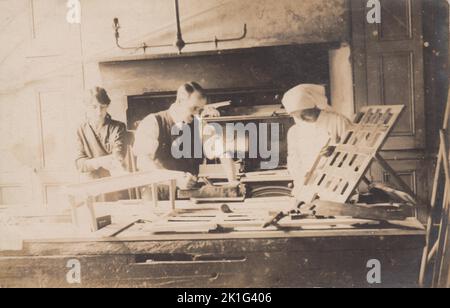 Fabricants de meubles, début du 20th siècle. Photographie d'un homme et de deux femmes qui font de la charpenterie et produisent des articles de meubles. L'homme utilise un plan de main sur un morceau de bois posé sur une grande table de travail. Une petite table et des cadres en bois avec sangle rembourrée sont également sur la table, avec des scies et d'autres outils de travail du bois. Une des femmes est vêtue d'un uniforme blanc, avec une tête couverte. Une gamme de cuisine peut être vue en arrière-plan. Banque D'Images