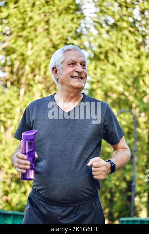 Un homme plus âgé de vêtements de sport tient une bouteille en plastique réutilisable debout ou fait du jogging dans le parc d'été, se soucier de la santé, faire de l'activité physique le matin. Sainement Banque D'Images