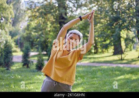 Femme asiatique active plus âgée dans les vêtements de sport regarder la caméra faire des exercices d'étirement dans le parc d'été avec des paumes repliées, faire de l'exercice de yoga ou d'entraînement Banque D'Images