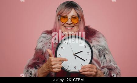 C'est votre temps. Portrait d'une femme âgée élégante montrant l'heure sur la montre, ok, pouce vers le haut, approuver, pointant le doigt vers l'appareil photo. Vieille grand-mère à l'intérieur studio tourné isolé sur fond rose Banque D'Images