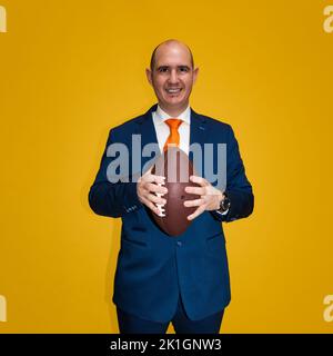 Un homme blanc casien vêtu d'un costume bleu, d'une chemise blanche et d'une cravate orange tenant une balle de football américain. L'arrière-plan est jaune. Banque D'Images