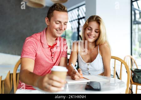 Des étudiants heureux assis dans la table de café de l'université et faisant des devoirs ou de la rédaction de plans. Une jeune femme et un homme passent du temps ensemble pendant le frein du café, en prenant des notes. Banque D'Images