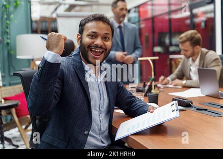 Un homme d'affaires indien heureux lors d'une réunion dans un bureau moderne et en regardant la caméra sur le fond de ses collègues Banque D'Images