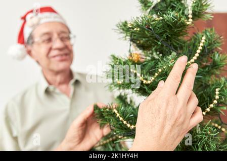 Couple hispanique senior décorant un arbre de Noël, appréciant les vacances ensemble. Mains de femmes organisant les décorations en avant-plan, Banque D'Images