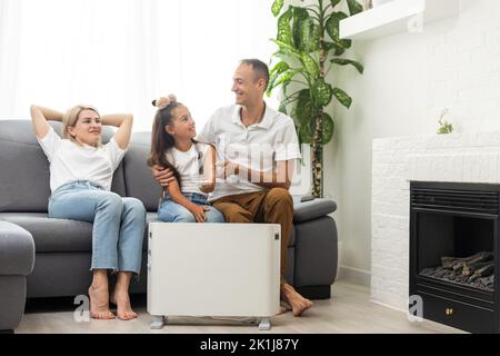 Chauffage électrique à la maison. Une jeune famille réchauffe ses mains gelées près d'un radiateur de chauffage Banque D'Images