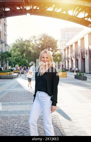 Gaie jeune femme blonde attrayante dans un pantalon blanc, regardant l'appareil photo. Pliez les genoux. Elle est debout sur un trottoir sur une rue ensoleillée. Banque D'Images