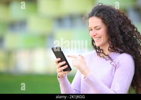 Une femme heureuse avec des marches de cheveux bouclés vérifier le contenu de smartphone dans la rue Banque D'Images