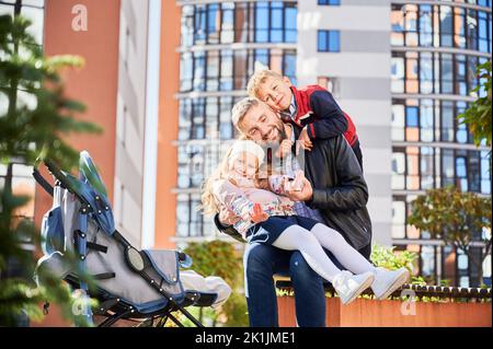 Famille gaie, portant des vêtements décontractés, se portant ensemble, tout en étant assis dans le parc avec un pram. Vue de face d'un père heureux assis sur le banc, souriant avec son fils et sa fille à l'extérieur. Concept de famille. Banque D'Images