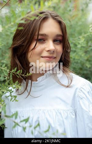 Portrait gros plan de jolie jeune fille en robe blanche avec plaisir debout parmi les branches de buissons avec des feuilles vertes, fond de la nature Banque D'Images