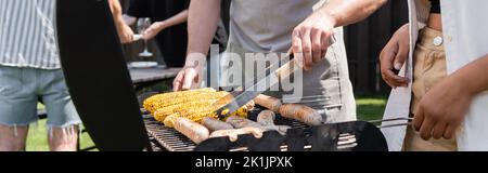 Vue rognée des amis interraciaux cuisant des saucisses et du maïs sur le grill dans l'arrière-cour, bannière Banque D'Images