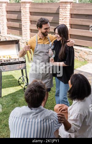 Homme souriant dans un tablier embrassant petite amie et tenant le vin près des amis multiethniques flous et de griller à l'extérieur Banque D'Images