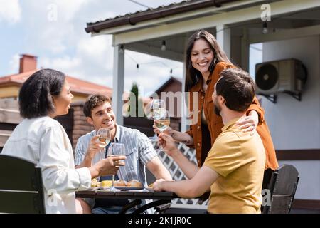 Jeune femme tenant du vin et embrassant l'homme près des amis interraciaux pendant le pique-nique dans l'arrière-cour Banque D'Images