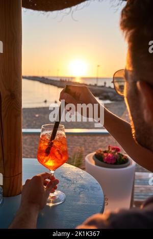 Un jeune homme qui apprécie et se détend en buvant une boisson fraîche lors d'une soirée européenne d'été tout en regardant le coucher de soleil méditerranéen dans un bar de plage. Style de vie Banque D'Images