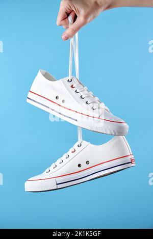 Sneakers de sport blanches pour femme accrochées à la main de femme avec lacets sur fond bleu, chaussures décontractées tendance, design minimaliste créatif Banque D'Images