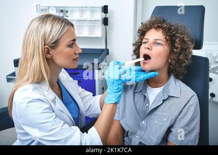 L'otolaryngologiste utilise une spatule d'inspection pour examiner la gorge de l'enfant mâle. Le jeune patient bouclés ouvre la bouche pendant le contrôle de la gorge Banque D'Images