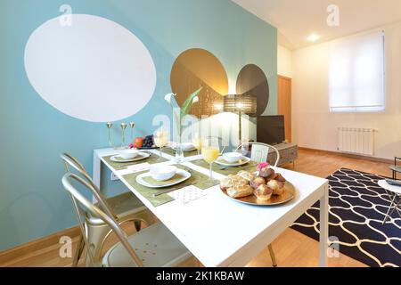 Appartement de vacances à la décoration éclectique avec une table à manger blanche avec un plateau de bonbons, un bol de fruits et un jus d'orange Banque D'Images