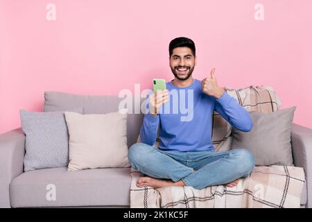 Photo de gai confiant gars habillé pull-over assis canapé montrant le pouce vers le haut dispositif moderne vide espace isolé couleur rose arrière-plan Banque D'Images