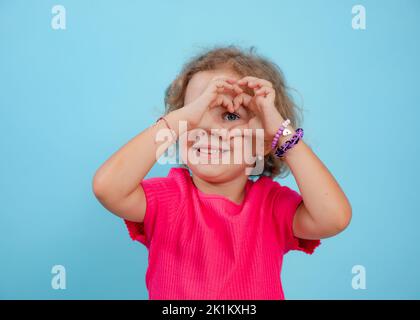 Petite fille avec des cheveux bouclés et équitables portant un T-shirt rose, regardant à travers les doigts en faisant la forme du coeur sur fond bleu. Banque D'Images