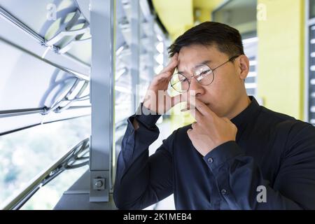 Homme d'affaires asiatique contrarié et triste dans la dépression fumer des cigarettes sur le balcon de l'immeuble de bureau, patron dans les lunettes bouleversé avec résultat avec de mauvaises pensées penser. Banque D'Images