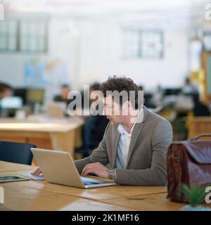 Il se distingue au bureau. Un homme mûr travaillant sur un ordinateur portable dans un grand bureau avec des collègues en arrière-plan. Banque D'Images