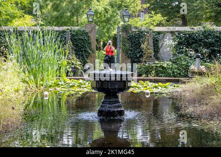 Étang avec une fontaine en pierre dans un jardin entouré de verdure, scène d'une femme photographe prenant une photo en arrière-plan, arbres verts luxuriants, soleil Banque D'Images