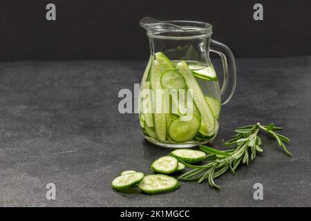 Eau perfusée au concombre dans une verseuse en verre. Branche de romarin sur table. Copier l'espace. Arrière-plan noir. Banque D'Images