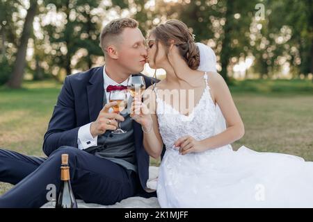 Portrait d'un merveilleux couple de mariage assis sur l'herbe verte dans le parc en été, tenant des verres de vin, embrassant. Amour. Banque D'Images