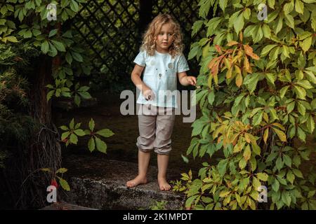 Portrait de la petite fille chérubique pieds nus debout sur de vieux escaliers dans un belvédère couvert de lierre en bois, jouant à cache-cache. Banque D'Images