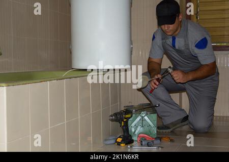 Plombier dans la salle de bains d'une maison qui, avec ses outils de travail, répare un flotteur d'un drain. Installation d'une chaudière dans la salle de bains. Banque D'Images