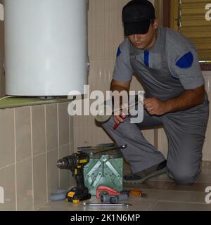 Plombier dans la salle de bains d'une maison qui, avec ses outils de travail, répare un flotteur d'un drain. Installation d'une chaudière dans la salle de bains. Banque D'Images