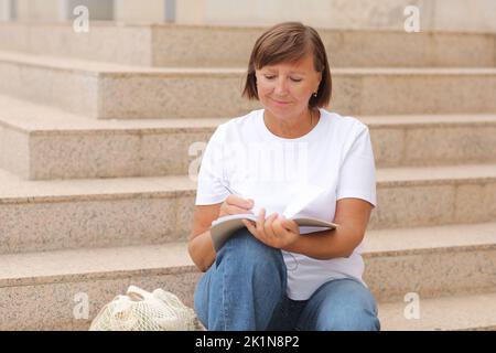 femme mature travaille à distance, l'enseignement supérieur pour les adultes, femme écrit et note dans un carnet assis sur les escaliers à l'extérieur, indépendant. Collège s Banque D'Images