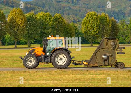 Un tracteur jaune avec une remorque de tondeuse à gazon industrielle conduit le long de l'aérodrome de l'aéroport, sur fond d'arbres Banque D'Images