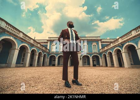 Un homme noir à barbe chauve majestueux, dans un costume et des lunettes rouge foncé à la coupe tendance, se trouve au centre d'une cour d'un anti bleu Banque D'Images