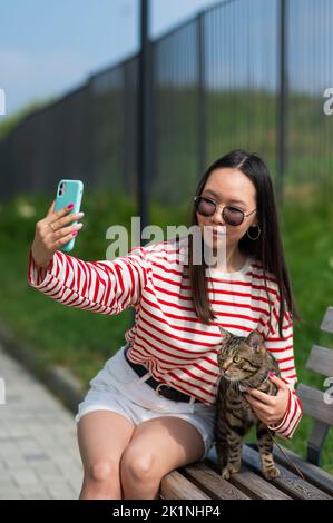 Une jeune femme s'assoit sur un banc avec un chat tabby et prend un selfie sur un smartphone à l'extérieur. Banque D'Images