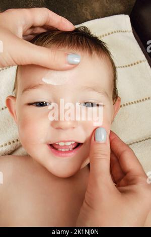 La mère sent de la crème sur le visage d'un bébé garçon heureux. Maman sourit de l'onguent sur la peau d'un enfant souriant. Enfant d'un an Banque D'Images