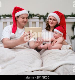 Homme heureux et femme avec enfant prendre des photos sur une tablette dans le lit décoré pour noël et nouvel an, couple de famille dans la chambre à la maison - Moscou, Russie, Decem Banque D'Images