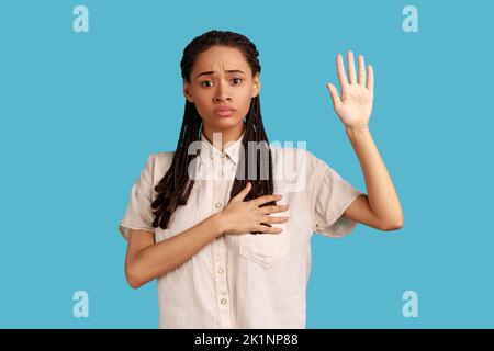 Je vous promets. Portrait d'une femme avec des dreadlocks faisant un geste d'assermentation et tenant le bras sur la poitrine, prêtant serment, juchant allégeance, portant une chemise blanche. Studio d'intérieur isolé sur fond bleu. Banque D'Images