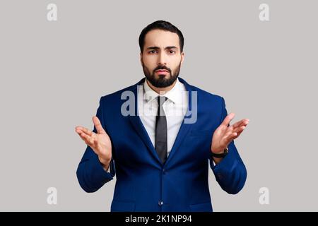 Portrait d'un homme barbu debout avec des mains levées et expression indignée confuse, demandant pourquoi, quelle raison, porter un costume de style officiel. Prise de vue en studio isolée sur fond gris. Banque D'Images