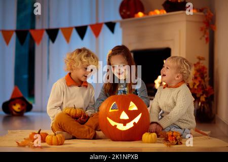Enfants sculptant de la citrouille pour Halloween à la maison assis à côté de la cheminée dans la salle de séjour décorée de lanternes et de feuilles d'automne. Trick or Treat. Banque D'Images