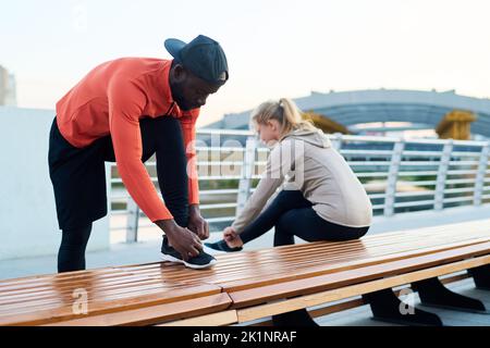 Jeune homme noir en casque d'activité se pliant au-dessus de la jambe et plié au genou tout en attachant la sneaker à la sportswoman assis sur le banc Banque D'Images