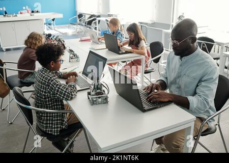 Groupe d'écoliers culturels créatifs et leur enseignant utilisant des ordinateurs portables en cours de robotique tout en étant assis devant l'un l'autre Banque D'Images