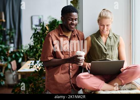 Joyeux jeune homme d'affaires avec une tasse de café regardant l'écran de l'ordinateur portable sur les genoux de son collègue tout en préparant la présentation Banque D'Images
