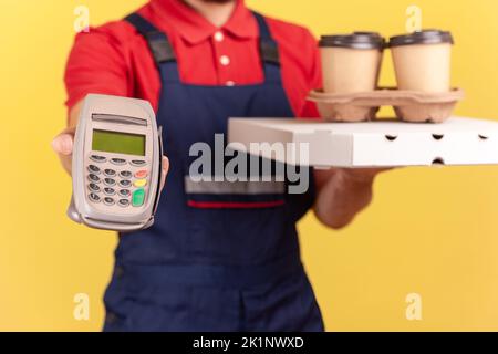 Un livreur anonyme debout avec une boîte à pizza et un café dans une tasse jetable et montrant le terminal de pos pour payer en ligne pour la livraison. Studio d'intérieur isolé sur fond jaune Banque D'Images